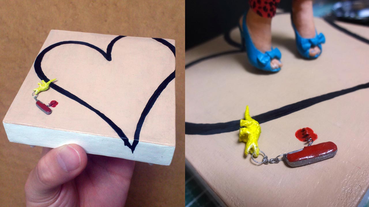 Two images in a close up of the finished base with tan color top with black line art of a heart and tiny swiss army knife corkscrew in a pool of blood attached to a mini yellow dinosaur at the mouth